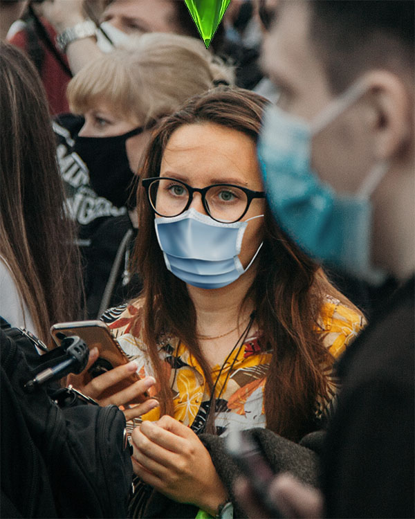A blank-faced, brown-haired young woman in a blue face mask, yellow patterned blouse, and black-framed glasses, holding a cell phone, looks off into the middle distance. Around her is a tightly-packed crowd of people, also in face masks, some with cameras, looking to the left at something we can't see. The woman is distant and disengaged in the middle of the crowd.