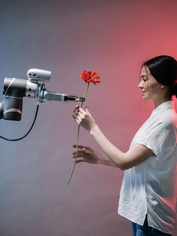 Against a smoky-looking background that shades from gray on the left to pink on the right, a woman (right) in a white, short-sleeved shirt and jeans, with her dark hair pulled into a low ponytail, accepts a long-stemmed red carnation from a gray and silver robot arm (left) — not the humanoid kind, just a robotic arm with a pincer hand and, for some reason, a flower. The woman is humanoid. It’s just the robot arm that isn’t.